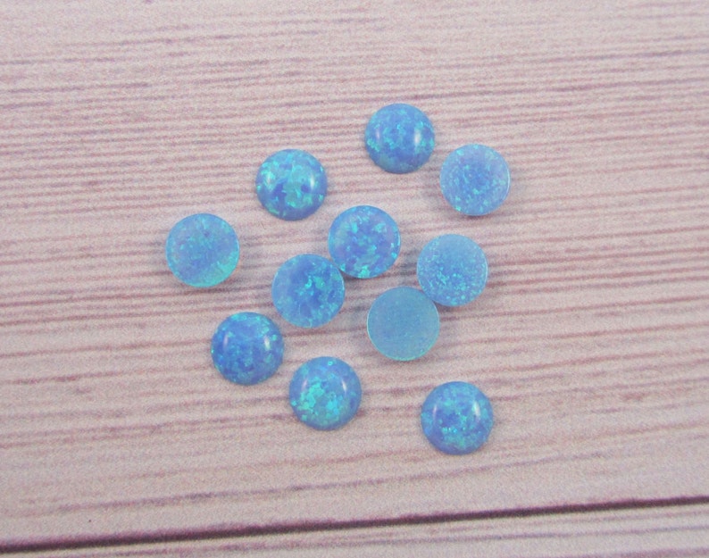 May include: A collection of 10 round, light blue opal cabochons. The cabochons are scattered on a light brown wooden surface.