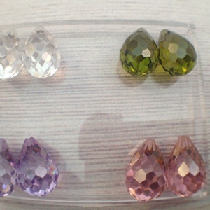 May include: Six teardrop-shaped crystal beads in a clear plastic container. Two beads are clear, two are green, and two are purple.