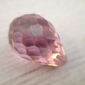 May include: A faceted, teardrop-shaped crystal bead in a pale pink color.