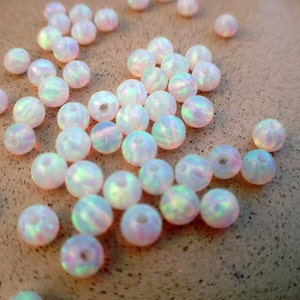 May include: A collection of iridescent white opal beads, each with a slightly different pattern and shimmer. The beads are round and smooth, and they are scattered on a light brown surface.
