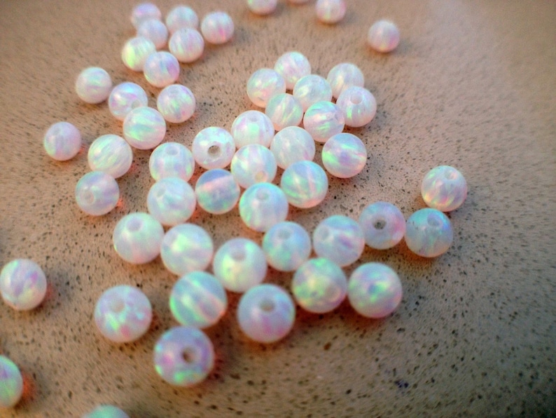 May include: A collection of iridescent white opal beads, each with a slightly different pattern and shimmer. The beads are round and smooth, and they are scattered on a light brown surface.