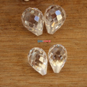 May include: Four clear faceted teardrop-shaped crystal beads. The beads are arranged in two pairs on a wooden surface.