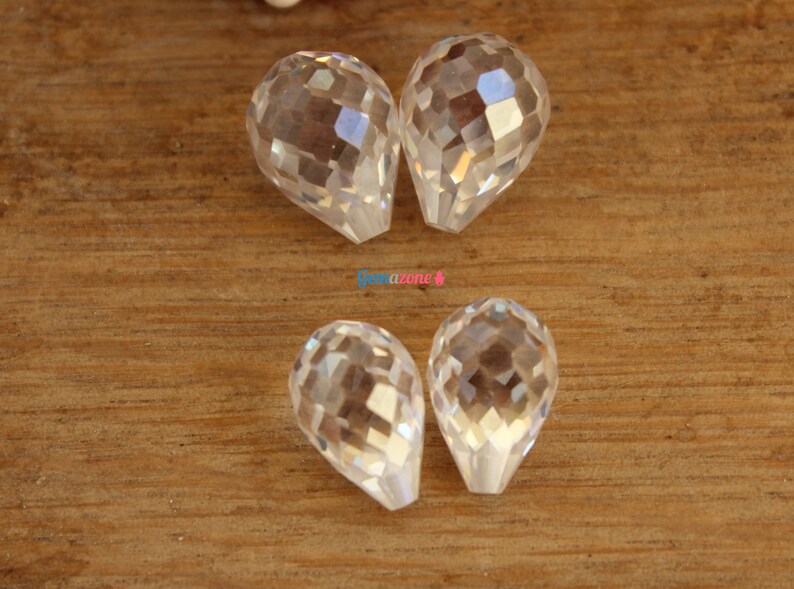 May include: Four clear faceted teardrop-shaped crystal beads. The beads are arranged in two pairs on a wooden surface.