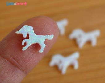 Könnte beinhalten: Ein kleiner, weiß schillernder Einhorn-Anhänger wird zwischen Fingern gehalten, mit weiteren im Hintergrund. Der Anhänger hat ein Horn, eine Mähne und einen Schwanz. Der Hintergrund ist eine verschwommene Holzoberfläche.