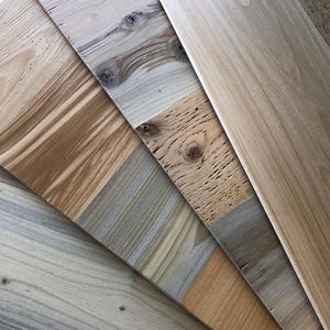 May include: An assortment of wooden planks in various shades of brown, beige, and gray. The planks display different grain patterns and textures, showcasing the natural variations of the wood. The arrangement suggests a display of wood samples or flooring options.