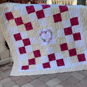 May include: A patchwork quilt featuring a checkerboard pattern of deep magenta and cream-colored fabric squares. The center square displays a floral wreath graphic surrounding the name Janice in cursive script with additional text below.