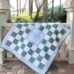 May include: A square quilt featuring a checkerboard pattern of sage green and white fabric squares. The centre square displays the name Calum in cursive script. The quilt is bordered by a thin floral print fabric and has intricate quilted stitching.