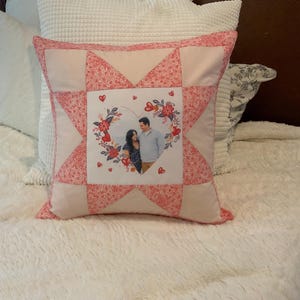 Puede incluir: Una almohada acolchada rosa y blanca con una foto en forma de corazón de una pareja frente a un diseño floral. La almohada tiene un patrón de estrella.
