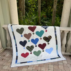Pode incluir: Um edredom branco com desenhos em forma de coração em várias cores e padrões, incluindo vermelho, azul, verde e camuflagem. O edredom tem uma borda estampada e é exibido em uma borda de pedra com um fundo natural.