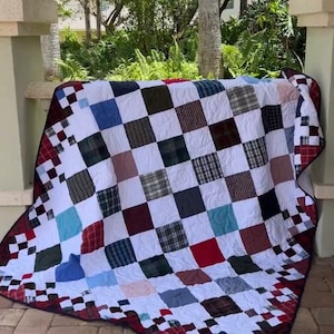 May include: A patchwork quilt with a checkered border. The quilt is made of various fabrics in shades of blue, red, white, and brown. The quilt is folded and displayed on a brick patio.