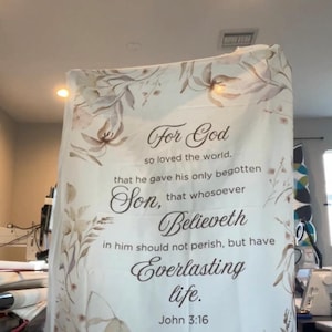 Puede incluir: Una manta blanca con detalles florales y el texto "Porque de tal manera amó Dios al mundo..." de Juan 3:16. La manta presenta una paleta de colores suaves y neutros con diseños florales marrones y beige.