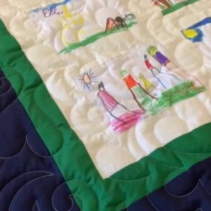 Op de afbeelding: Handgemaakte quilt met kindertekeningen. De witte quilt heeft een groene rand en een blauwe achterkant met een golfpatroon. De tekeningen zijn kleurrijk en tonen verschillende scènes, waaronder mensen en landschappen.