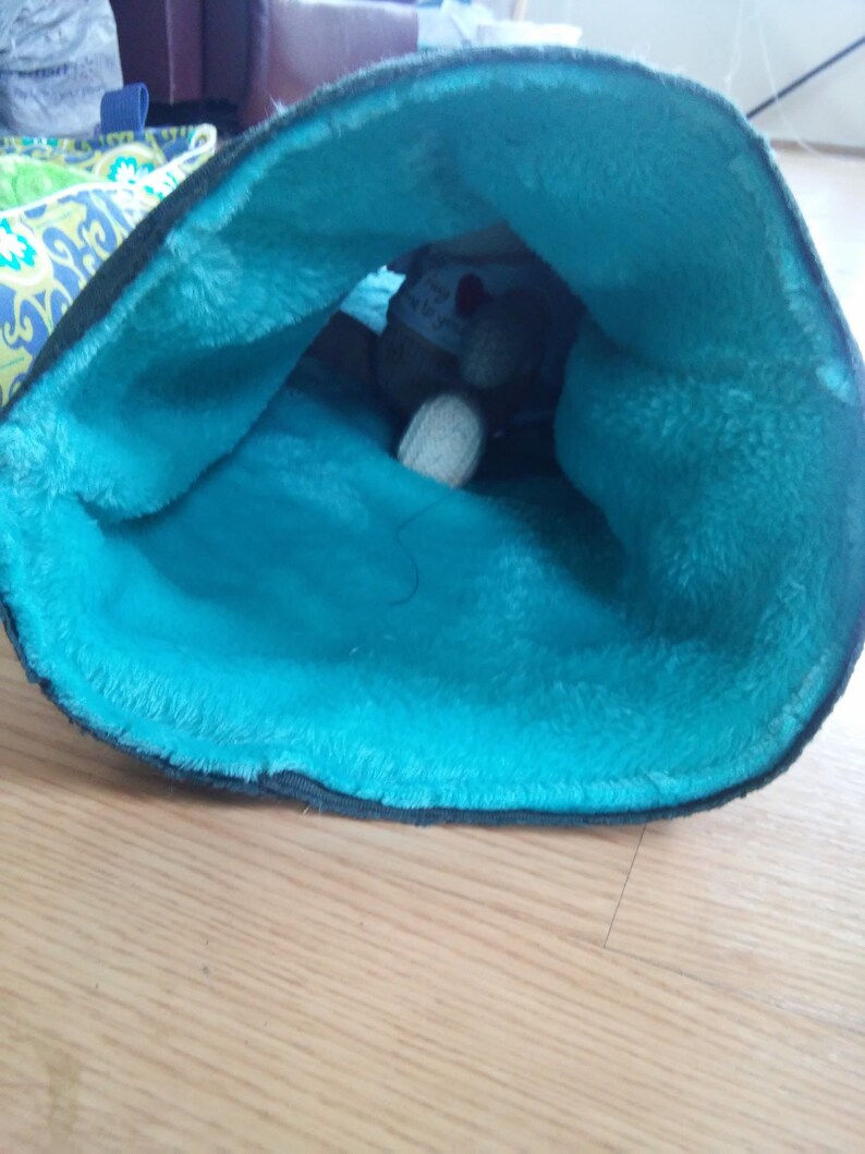 May include: A teal blue fabric-lined pouch with a stuffed animal inside. The pouch is made of a soft, fuzzy material.