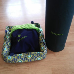 May include: A blue and green pet bed with a navy blue blanket inside. The blanket has the name "Sampson" embroidered on it. A black cylindrical object with the name "Sampson" embroidered on it is in the background.