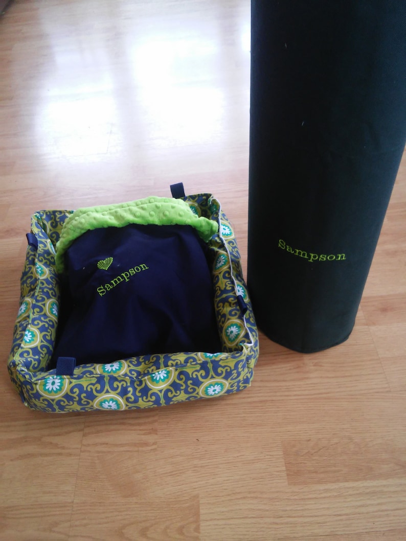 May include: A blue and green pet bed with a navy blue blanket inside. The blanket has the name "Sampson" embroidered on it. A black cylindrical object with the name "Sampson" embroidered on it is in the background.