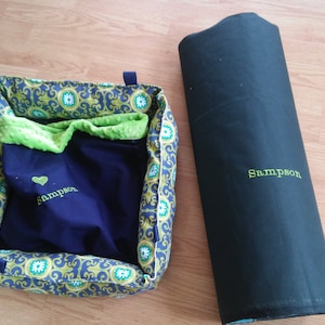 May include: A green and blue patterned pet bed with a removable green fleece liner. The bed is embroidered with the name "Sampson" and a heart. A black fabric roll with the name "Sampson" embroidered on it is also shown.