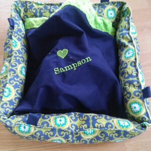 May include: A blue and green pet bed with a green and white patterned fabric. The bed has a blue blanket inside with a heart and the name "Sampson" embroidered on it.