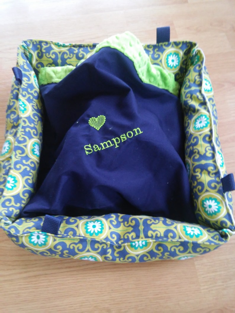 May include: A blue and green pet bed with a green and white patterned fabric. The bed has a blue blanket inside with a heart and the name "Sampson" embroidered on it.