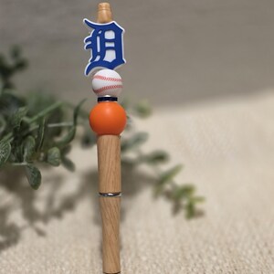 May include: A wooden pen with a baseball theme. The pen features a blue and white Detroit Tigers logo, a baseball, and an orange sphere. The pen has a silver band and a clear plastic stand.