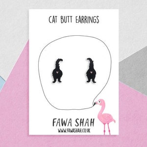 Op de afbeelding: Een witte kaart met zwarte tekst die "Cat Butt Earrings" en "Fawa Shah" met een websiteadres zegt. De kaart toont een zwart silhouet van twee kattenbillen en een roze flamingo.