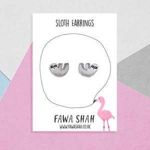 Könnte beinhalten: Eine weiße Karte mit dem Text "Sloth Earrings" und "FAWA SHAH" mit einer Illustration eines rosa Flamingos. Die Karte zeigt zwei silberne Faultier-Ohrringe.