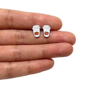 Op de afbeelding: Twee witte oorbellen in de vorm van koffiebekers met zwarte contouren en oranje pompoenontwerpen. De oorbellen zijn klein, waarschijnlijk gemaakt van hout of een vergelijkbaar materiaal, en worden in een hand tegen een witte achtergrond gehouden.
