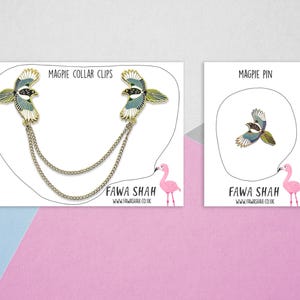 Puede incluir: Dos clips de collar de p&aacute;jaro urraca dorados con una cadena y un solo pin de p&aacute;jaro urraca. Los pines est&aacute;n en tarjetas blancas con flamencos rosas y el texto "Magpie Collar Clips" y "Magpie Pin". El texto "Fawa Shah" y una direcci&oacute;n de sitio web tambi&eacute;n est&aacute;n en las tarjetas.