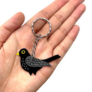 Puede incluir: Un llavero de pájaro negro con pico y ojo amarillos. El pájaro tiene detalles de alas grises y está sujeto a un llavero plateado. El llavero se sostiene en una mano.