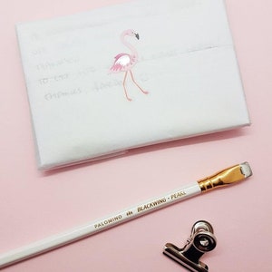 Puede incluir: Un sobre blanco con una pegatina de flamenco rosa. Un l&aacute;piz blanco con letras doradas que dicen "PALOMINO BLACKWING PEARL" est&aacute; sobre una superficie rosa. Un clip de carpeta plateado tambi&eacute;n est&aacute; sobre la superficie.