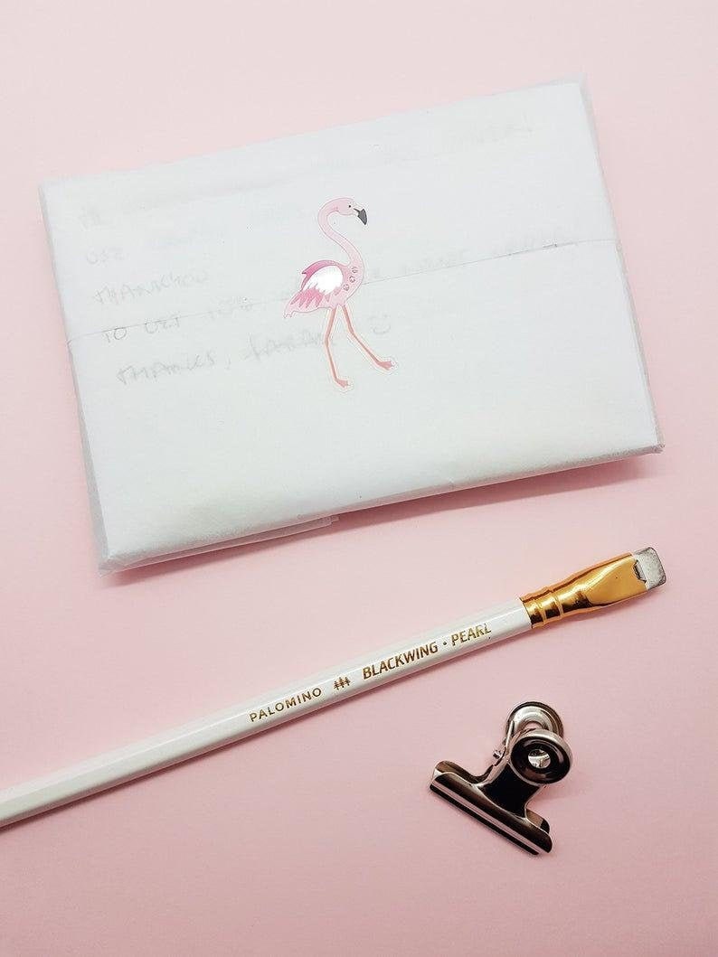 Puede incluir: Un sobre blanco con una pegatina de flamenco rosa. Un l&aacute;piz blanco con letras doradas que dicen "PALOMINO BLACKWING PEARL" est&aacute; sobre una superficie rosa. Un clip de carpeta plateado tambi&eacute;n est&aacute; sobre la superficie.
