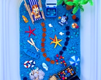May include: A sensory bin with blue dyed rice, featuring miniature beach-themed toys. Includes a miniature beach chair, toy boats, starfish, seashells, a treasure chest, and a palm tree. The bin is in a white plastic container.
