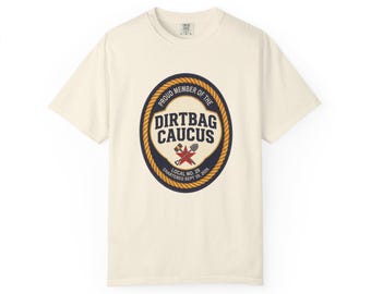 Könnte beinhalten: Ein cremefarbenes Kurzarm-T-Shirt mit einem zentralen ovalen Emblem mit Seilrand. Die Grafik enthält den Text "PROUD MEMBER OF THE DIRTBAG CAUCUS", "LOCAL NO. 29" und "CHARTERED SEPT 29, 2026" sowie einen roten Stern, einen Hammer und eine Schaufel.