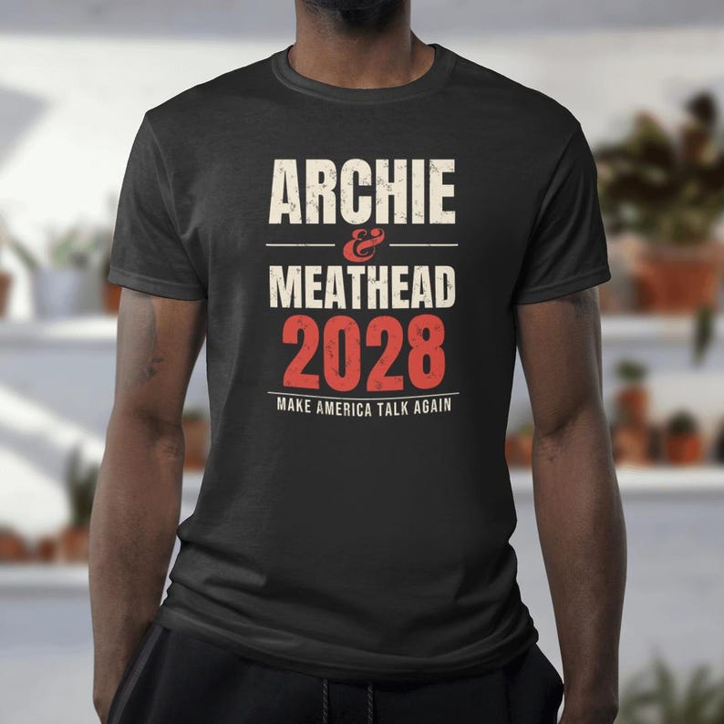 May include: A black short-sleeved t-shirt featuring white and red distressed-style text that reads ARCHIE & MEATHEAD 2028 MAKE AMERICA TALK AGAIN. The design is centered on the chest with a crew neckline and a standard fit.