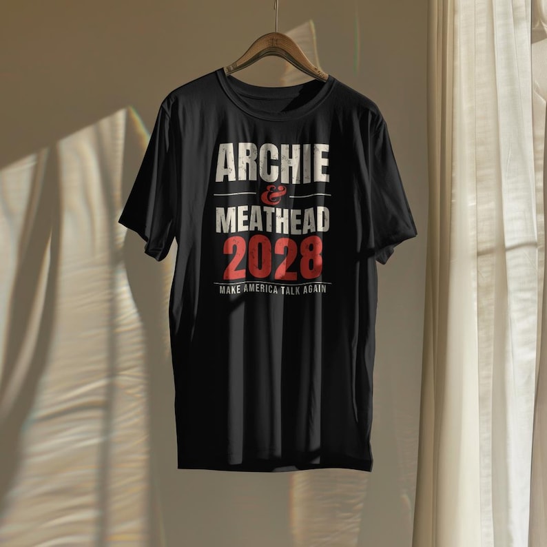May include: A black short-sleeved t-shirt featuring white and red distressed text that reads ARCHIE & MEATHEAD 2028 and MAKE AMERICA TALK AGAIN. The shirt is displayed on a wooden hanger against a neutral background with soft lighting.