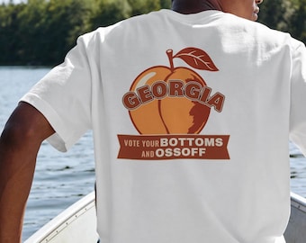May include: A white short-sleeve t-shirt featuring a graphic on the back of a peach with a leaf. The text GEORGIA is arched over the peach, and a banner below reads VOTE YOUR BOTTOMS AND OSSOFF in bold, dark orange lettering.