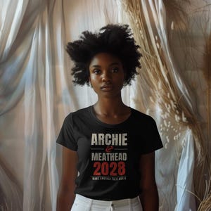 May include: A black short-sleeved t-shirt featuring the white and red text ARCHIE & MEATHEAD 2028 and the smaller text MAKE AMERICA TALK AGAIN printed on the front. The shirt has a crew neckline and a standard fit.
