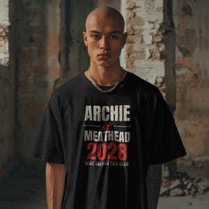 May include: A black short-sleeved t-shirt featuring white and red text that reads ARCHIE & MEATHEAD 2028 MAKE AMERICA TALK AGAIN. The shirt has a crew neckline and is worn with a silver-toned chain necklace.