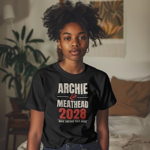 May include: A black short-sleeved t-shirt featuring white and red text that reads ARCHIE & MEATHEAD 2028 MAKE AMERICA TALK AGAIN. The design is centered on the chest with a distressed, vintage-style font and bold red numbers.
