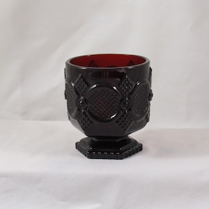 May include: A dark red, footed glass goblet. The goblet has a patterned design with a textured surface. The interior of the goblet is a deep red color. The base is a square shape.