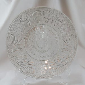 May include: Clear glass plate with embossed floral and scrollwork design. The plate features a central floral pattern and a decorative border. The plate is displayed on a clear plastic stand. A decorative serving piece.