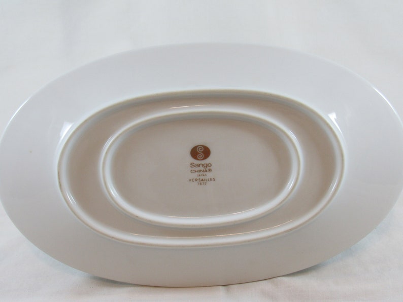 May include: A white oval-shaped serving dish with a raised rim. The bottom of the dish has a brown logo that reads "Sango China" and "Versailles 18-12".