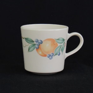 May include: A white ceramic mug with a fruit design. The design features a peach, green leaves, and blue berries.