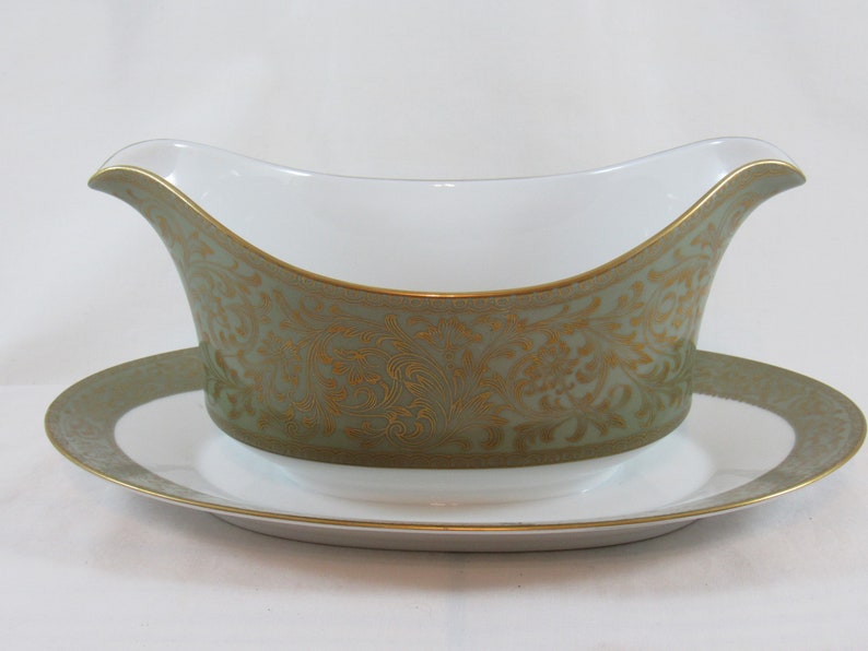 May include: A white porcelain gravy boat with a green and gold floral pattern. The boat has a matching saucer.