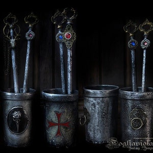 Può includere: Quattro contenitori metallici con disegni decorativi contengono penne metalliche ornate con punte gioiello. I contenitori sono dipinti in una finitura nera invecchiata. I disegni sui contenitori includono un teschio, una croce, un motivo floreale e un cerchio con un numero.