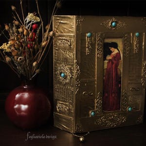 May include: A gold-colored book with intricate designs and turquoise gemstones. The book has a picture of a woman in a red dress on the front cover.