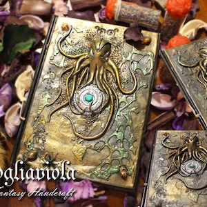 May include: A handmade journal with a gold and green cover featuring a detailed embossed design of an octopus with a jeweled eye. The journal is titled "Fogliaviola Fantasy Handcraft".