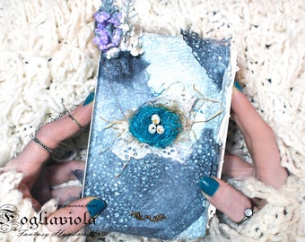 May include: A small, blue and white journal with a bird's nest and eggs on the cover. The journal has a distressed, vintage look and is decorated with lace and flowers. The text "Fogliaviola.com Fantasy Handmade" is printed on the bottom of the cover.