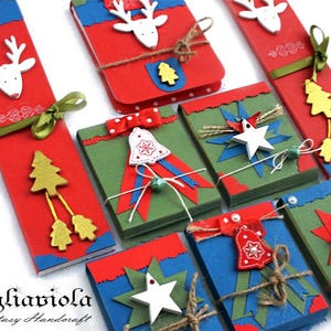 Può includere: Una collezione di confezioni regalo e segnalibri a tema natalizio fatti a mano. Le scatole e i segnalibri sono rossi, verdi e blu, decorati con renne bianche, stelle, campane e alberi di Natale. Alcuni sono legati con spago e nastri.