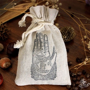 Può includere: Un piccolo sacchetto di lino bianco con coulisse e una stampa in bianco e nero di una mano con linee e simboli. Il sacchetto è su una superficie di legno con vari oggetti naturali.