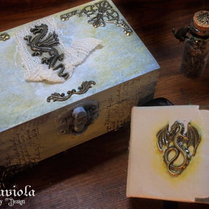 May include: A decorative wooden box with a light blue and gold finish. The box features a metal dragon design and a lace overlay. The box is closed and has a metal latch. A small glass bottle with a cork top and a metal dragon design is also pictured.